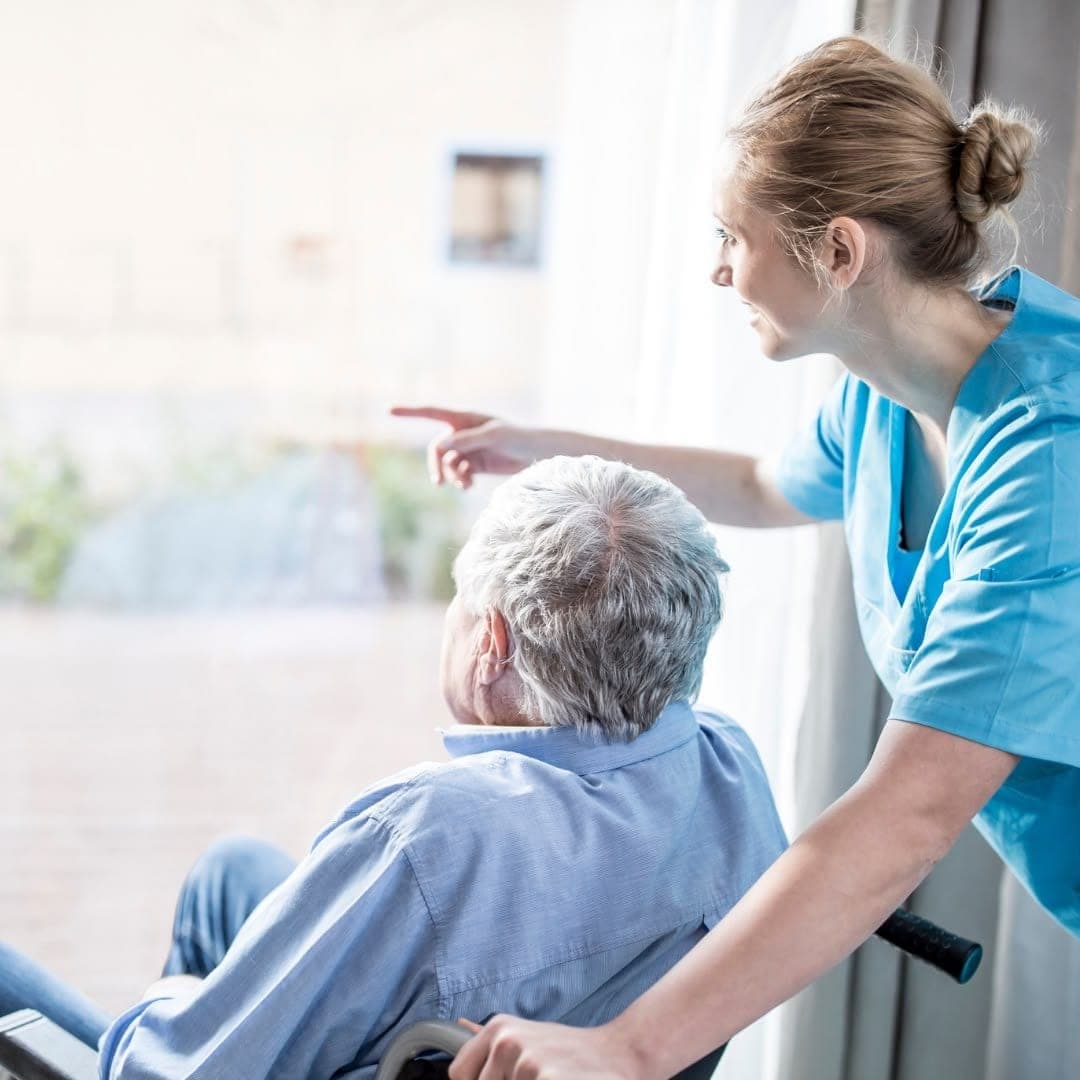 A carer in uniform beside an older man in a wheelchair, pointing out of a window